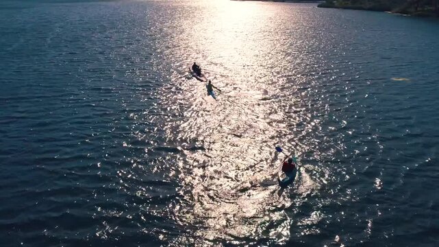 Drone shot of kayakers and rowers moving across sunlit lake water with bright reflections and ripples. Scenic outdoor sport scene showing teamwork, training, freedom and nature.