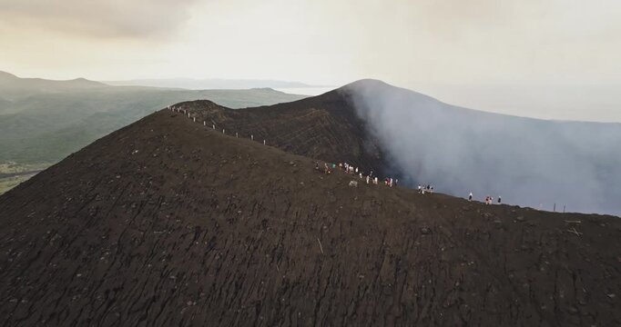Tourists standing on dark ash rim of Mount Yasur volcano, looking down into active crater emitting large plumes of white smoke on Tanna Island, Vanuatu. Travel, explore, adventure. Aerial drone shot
