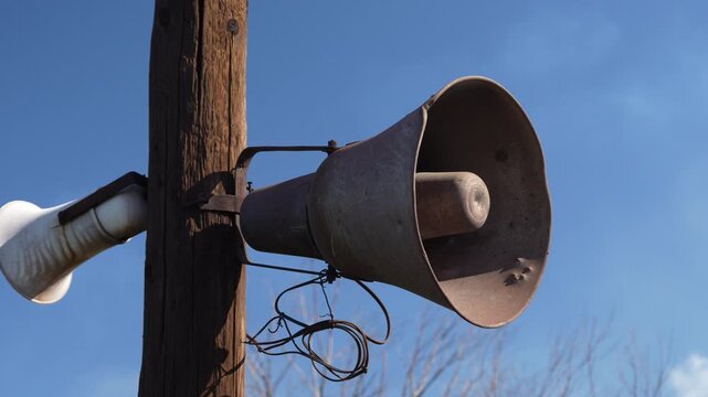 Vintage aluminum loudspeaker mounted on a wooden utility pole against clear blue sky. Static shot of retro public announcement equipment outdoors.