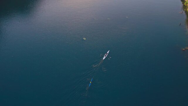 Top-down drone shot of a kayaker and rowing boat moving across a deep blue lake with wide copy space. Minimal outdoor sports scene showing calm water, training, teamwork and nature.