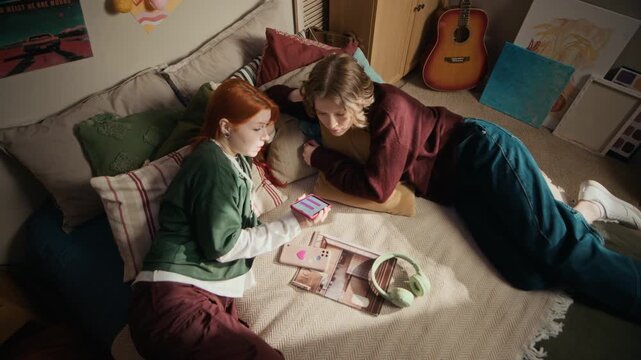 Top view of two young Caucasian women relaxing on bed while browsing smartphone together in cozy bedroom apartment