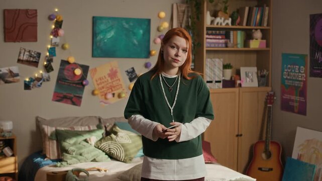 Medium portrait of Caucasian teenage girl with red hair wearing green long sleeve standing in stylish cozy bedroom and holding stack of polaroid photos while looking at camera, smiling shyly
