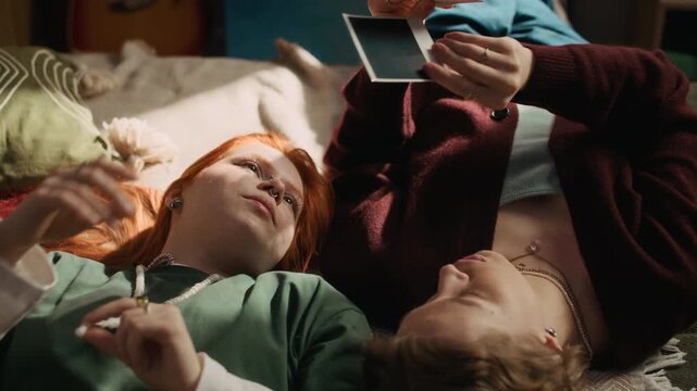 Female friends relaxing on bed and viewing instant printed photos together in cozy home setting