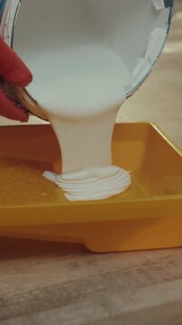 White paint flows steadily into yellow tray now, Gloved hand guides viscous white paint into yellow tray carefully