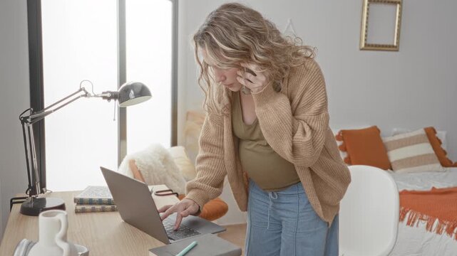 Pregnant woman holding phone to ear and typing on laptop at bedroom desk with lamp and cushions; focused multitasking.