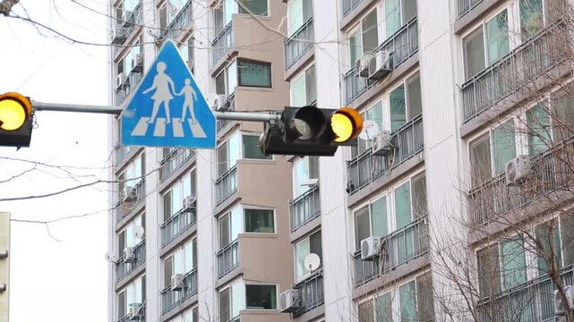아파트 단지 신호등 표지판 풍경 동해: 고층 아파트 외벽을 배경으로 도로 위의 보행자 표지판과 신호등을 로우 앵글 구도로 촬영한 도시 풍경 영상입니다. 실외기가 설치된 주거 건물과 교통 안전 시설물을 와이드 숏으로 담았으며, 한국 특유의 아파트 주거 환경, 주택가 일상, 어린이 보호 구역 및 사색적인 드라마 인서트 숏 콘셉트를 상징합니다.