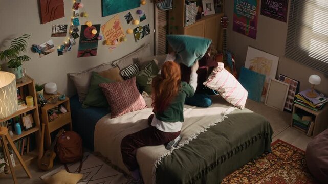 Top view shot of two young Gen Z female friends laughing and having playful pillow fight on bed in cozy bedroom at home