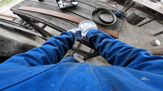 Worker in blue protective overalls and gloves opens a can of brown varnish or wood stain. First person view (POV). The craftsman is preparing to paint a wooden part, around controlled chaos