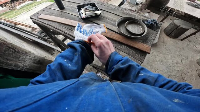 Worker in blue protective overalls and gloves opens a can of brown varnish or wood stain. First person view (POV). The craftsman is preparing to paint a wooden part, around controlled chaos