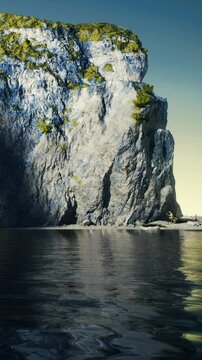 A cliff stands beside still water. The rock face is tall and rugged. The water reflects the cliff and the sky. Green plants grow on top of the cliff in bright sunlight.