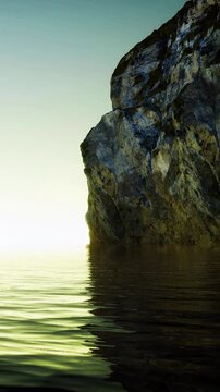 Early morning light shines on still water by a rocky shore. The sun rises in the background, creating a soft glow. The rock is covered in moss, enhancing the natural beauty of the scene.