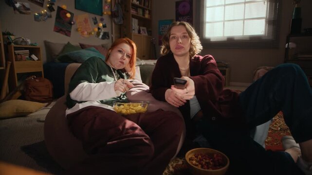 Stab shot of two young Caucasian female friends watching TV and sharing snacks and conversation while spending relaxing time together in bright cozy bedroom