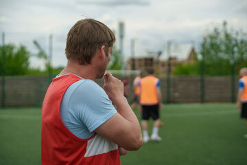Man in a red and white vest standing on a soccer field during a team break outdoors, football player coach, casual sport discussion. © neteli