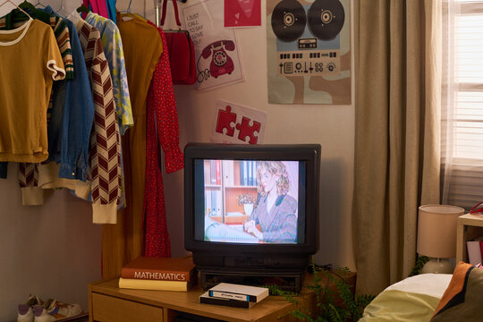 Wide shot of retro bedroom displaying vintage television, VHS player, hanging clothes, nostalgic posters, evoking home entertainment and 1980s lifestyle. For editorial, nostalgia marketing