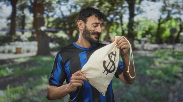 Man in soccer jersey holding a money bag with dollar sign, smiling and inspecting its drawstring while standing in forest; satisfaction.