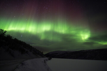 Glowing green northern lights band above a snowy arctic road and dark silhouette of hills at night © Benjamin