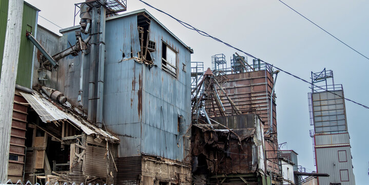 Buildings at an abandoned industrial site stand in disrepair. Metal and wood show signs of wear. Clouds hang over the area. The structures hint at past activity and leave a somber feeling.