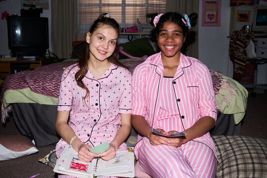 Portrait of Caucasian teenager and Black teenager sitting together, holding notes and smiling during sleepover activity, supporting friendship and youth lifestyle marketing