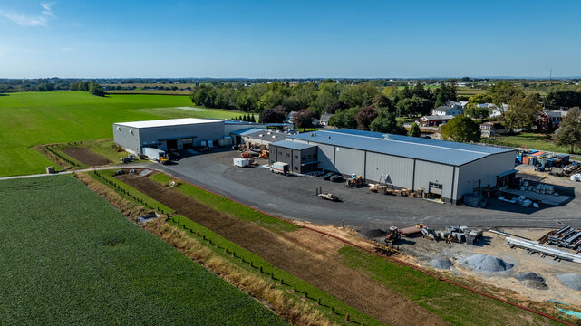 Large farm buildings are seen in a rural setting. Green fields surround the area, and equipment is parked nearby. The sky is clear and bright.