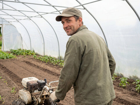 Farmer tilling soil with rotary cultivator inside polytunnel greenhouse