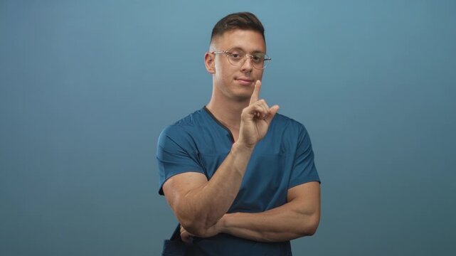 Man doctor wearing blue scrubs and glasses, young blond handsome points right index finger with folded arms in studio neutral backdrop; confidence professionalism.