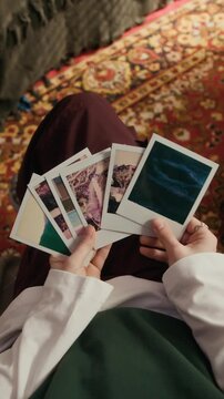 Vertical top view shot of teenage girl relaxing in armchair while holding instant photo prints in cozy bedroom with nostalgic mood