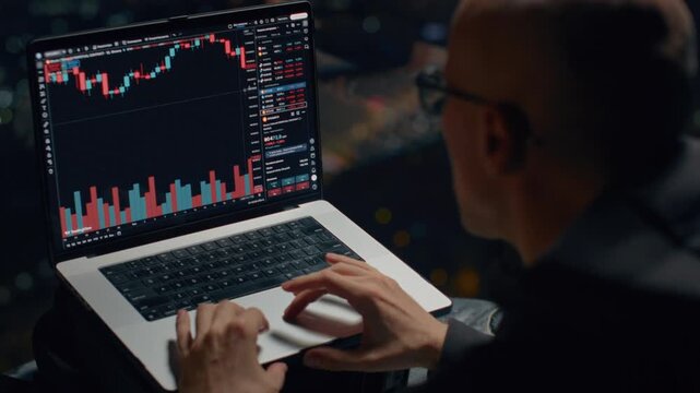 Bald man trader trades on stock exchange on laptop at home at night, lives in a skyscraper, a night city. Male investor analyzes stock market and cryptocurrencies. Apartment in downtown. BTC Ethereum