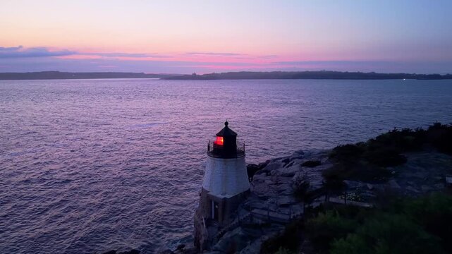 Castle Hill Lighthouse at sunset aerial video, a scenic drone shot of the historic beacon illuminated on a rocky cliff point during a vibrant twilight in Newport, Rhode Island.