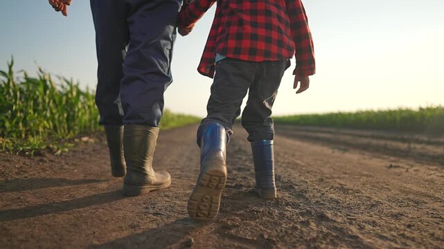 Farmer and boy walking dirt road at sunset. Man leads child with gentle care near summer field. Boots crush warm ground. Walking side by side builds bond. Road stretches far under soft evening sky.