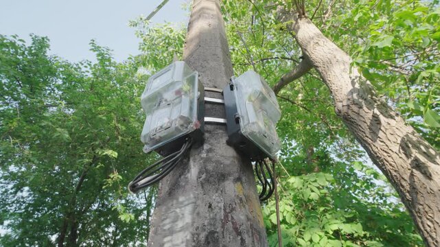 Low-Angle Bottom-Up View: Electric Meters in Plastic Cases Mounted on Concrete Utility Pole | Outdoor Power Consumption Devices with Cables in Green Trees | Rural Electricity Infrastructure