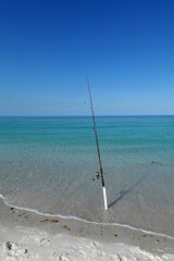 Fishing poles stand upright in the sand along a serene beach. The clear turquoise water gently laps at the shore under a cloudless blue sky.  © Gene