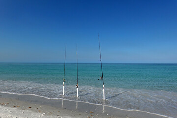 Fishing poles stand upright in the sand along a serene beach. The clear turquoise water gently laps at the shore under a cloudless blue sky.  © Gene