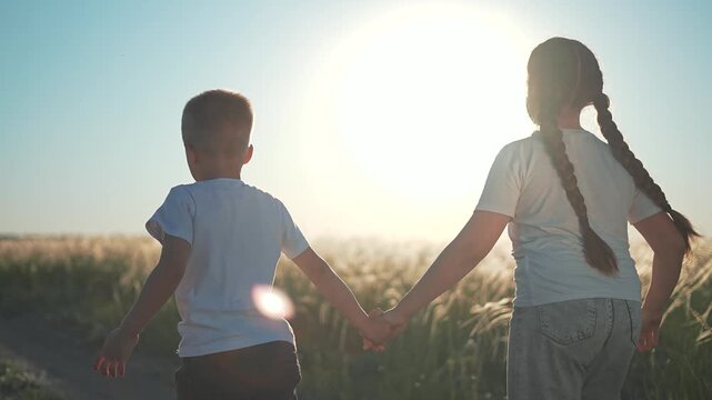 Girl and boy child holding hand move along field path under bright sunset. Smiling sibling together step carefully. Child faces glow warm light. Brother sister love walking field hand in hand.