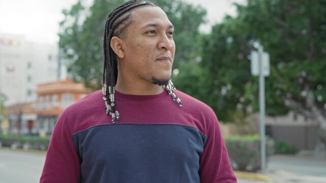 Man with braided hair and beaded ends smiles and looks to the right while standing on a street near a building and trees; optimism.