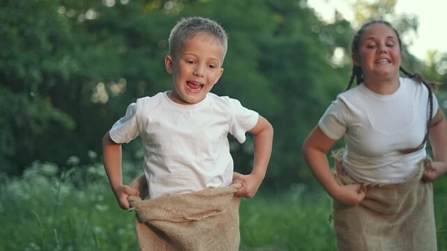 Boy jumps in front during summer game. Girl smiling behind joins fun outdoors. Jumping kid leads competition. Active children in sack race play together. Boy and girl enjoy summer jumping game.