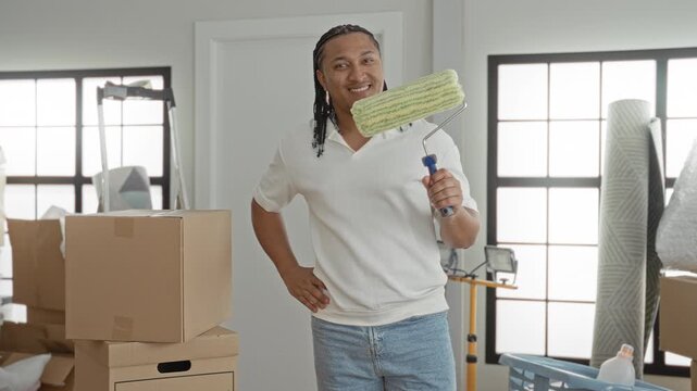 Man holding paint roller with right hand and hand on hip among moving boxes and rolled rugs inside a building; home pride.