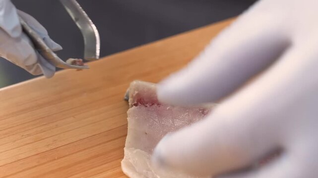 A static up-close shot captures a professional chef, wearing white gloves, carefully removing fish bones from a raw, skinless fish fillet on a wooden cutting board.