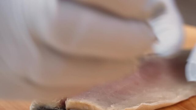 detailed close-up shot of a professional chef wearing white protective gloves, carefully preparing a fresh fish fillet on a wooden cutting board. The footage highlights the precision involved in clean