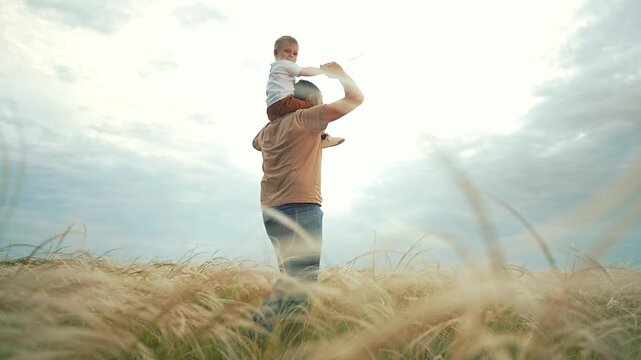 Kid sits on father shoulders while walking. Parent bonding with child in nature. Kid smiles with father. Boy enjoys walking with parent. Kid bonding in open air. Father and kid walk together.