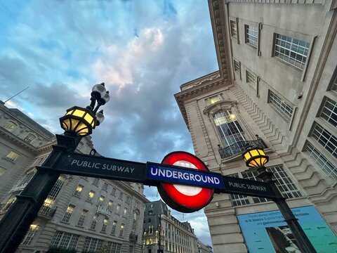 Wide low-angle view of an illuminated London Underground public subway sign and vintage street lamps at twilight surrounded by grand city architecture