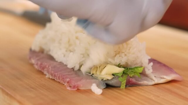 close-up shot of a professional sushi chef's hands carefully assembling Saba Bo-zushi. The footage captures the process of spreading vinegared sushi rice over a fresh, vinegar-cured mackerel fillet (S