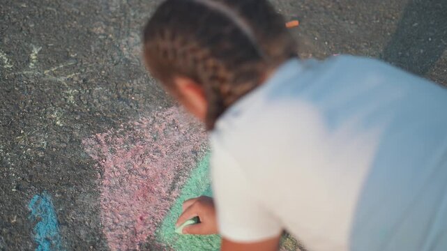 girl bends low, chalk in hand, drawing on street. Chalk dust floats in summer air, child smiling in outdoor play. kid loves art, drawing again with chalk, summer colors bright on street in happy art.
