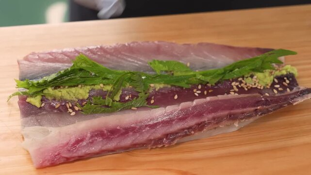 Close-up footage of a sushi chef wearing blue gloves meticulously assembling a traditional Japanese dish. The chef is seen placing fresh green shiso leaves and wasabi onto a large fillet of seared bon