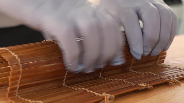 Close-up footage of a professional chef wearing hygienic gloves, skillfully using a traditional bamboo rolling mat (makisu) and plastic wrap to shape a fresh sushi roll.