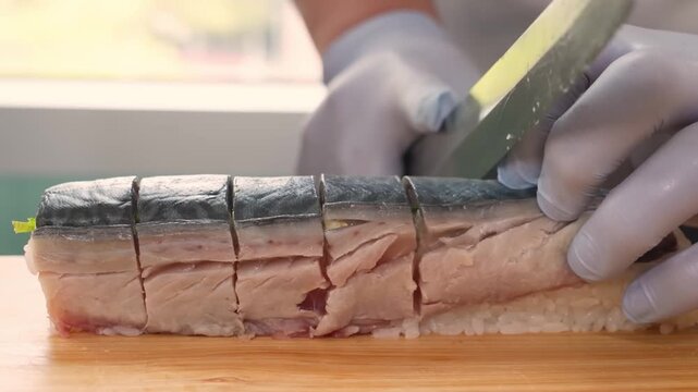 Close-up of a professional sushi chef&rsquo;s hands wearing protective gloves, expertly slicing a long roll of traditional Kyoto-style pressed mackerel sushi (Saba-zushi). The footage highlights the precisi