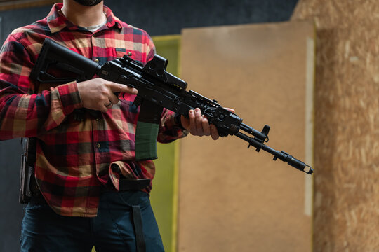 Man in plaid shirt holding tactical rifle with collimator sight at indoor shooting range training