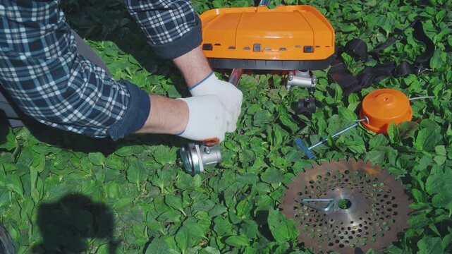 Man in a plaid shirt works on assembling gardening tool or trimmer components while kneeling on lush green grass, with various parts and tools arranged nearby in the outdoor environment