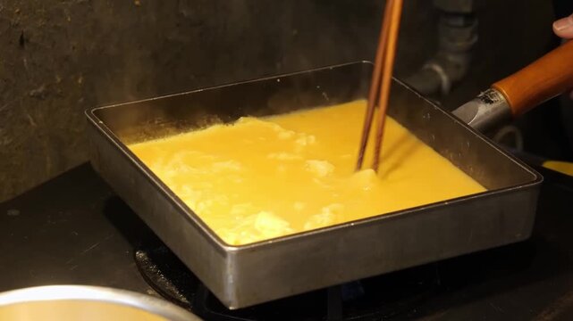 A close-up shot of the traditional process of making Tamagoyaki, a Japanese rolled omelet. The footage shows a thin layer of seasoned egg mixture being cooked in a professional rectangular Tamagoyaki 