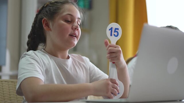 Girl holds number flashcard during online learning session with laptop at home. Student practices math using educational flashcard. Child learns numbers through digital education at desk.