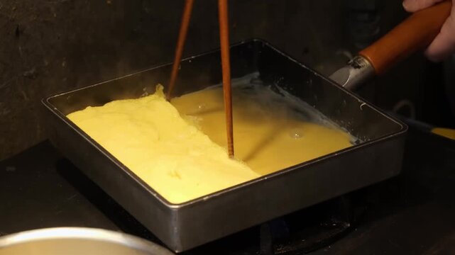 A close-up shot of the authentic process of making Japanese Tamagoyaki. Liquid egg mixture is poured into a traditional square tamagoyaki pan and carefully rolled in layers. This classic side dish is 